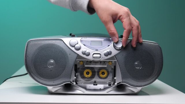 Hand adjusting the volume on a classic silver boombox, featuring a visible cassette tape, against a vibrant turquoise background, symbolizing retro music and sound technology