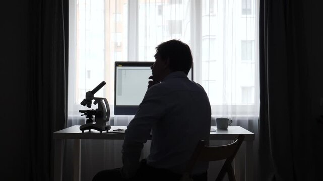 Scientist silhouette working on a desktop computer with a microscope on the desk, conducting research and analysis in a modern laboratory or home office setting