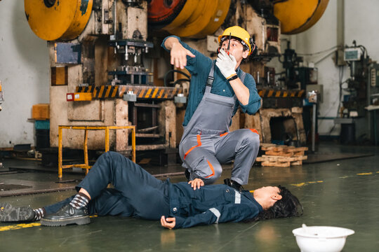 Industrial worker calling emergency service beside injured colleague on factory floor during workplace accident and safety response training