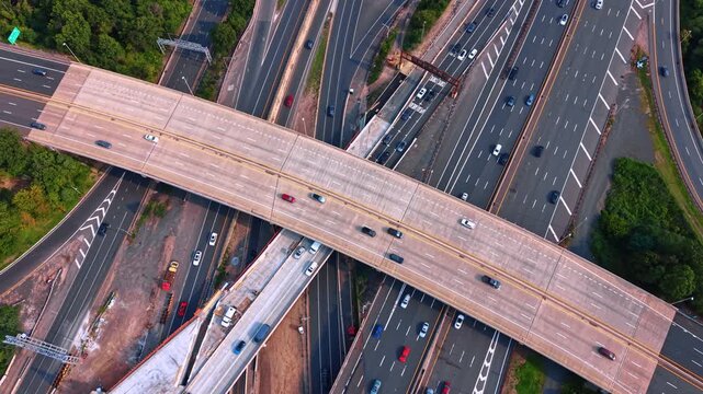 Footage over a massive multi-level highway interchange bustling with moderate vehicle traffic. A wide concrete overpass bridging over a dense network of multi-lane expressways running underneath.