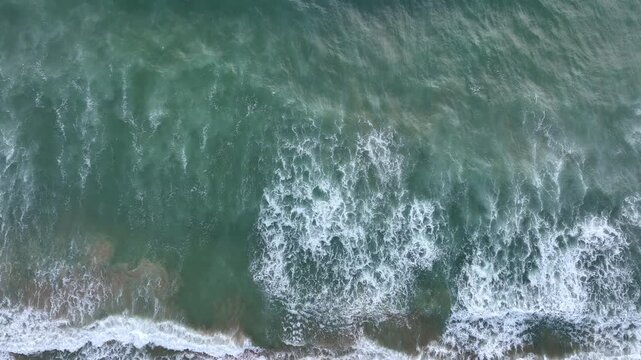 Foamy waves crashing from above. Aerial drone view of sea foam. Ocean foam moving on shoreline. Drone captures foamy surf. White foam spreading on beach from drone.