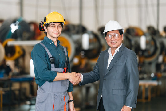 Industrial worker and business manager shaking hands inside factory, wearing safety helmet and workwear, expressing trust, teamwork, and professional cooperation in manufacturing