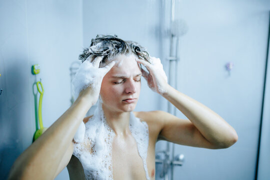 Teen boy holding soapy hands on head under the warm bathroom shower.