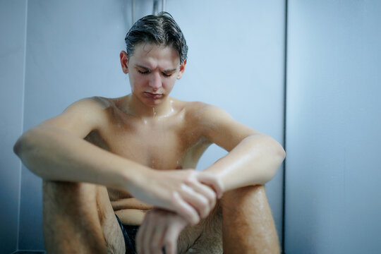 Stressed young guy resting on the wet bathroom cabin floor indoors.