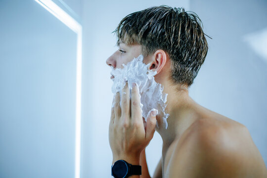 Young adolescent guy applying shaving foam on face facing bathroom mirror.