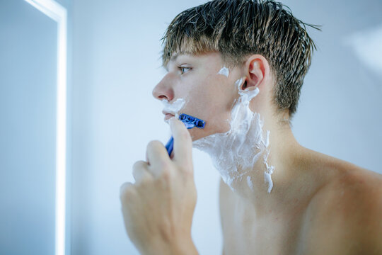 Teenager shaving his face with a blue razor and white foam in the bathroom.