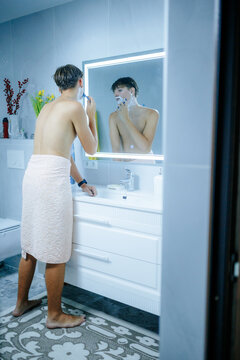 Young guy removing facial hair using sharp razor before bathroom mirror.