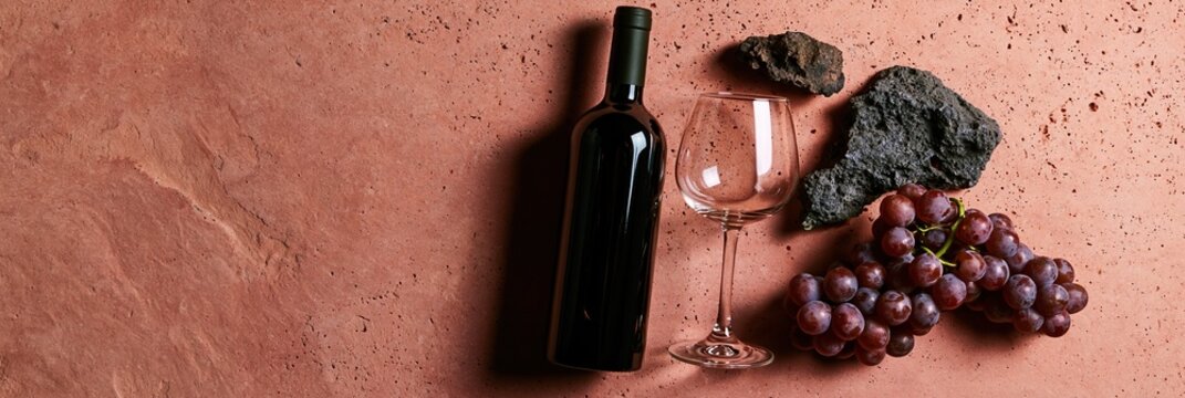 Dark wine bottle and clear glass sit with red grapes and volcanic stone on warm pink tuff surface in controlled lighting.