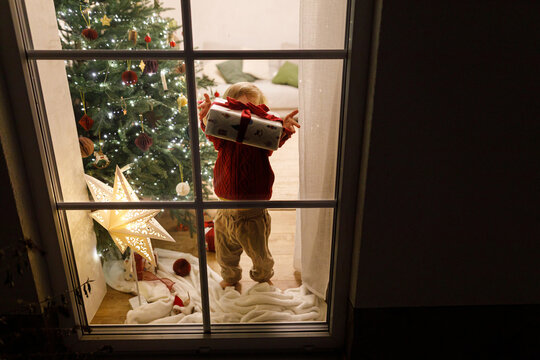 Toddler holding wrapped Christmas present behind window near illuminated tree. Holiday excitement, festive gift giving, cozy home winter celebration. Merry Christmas