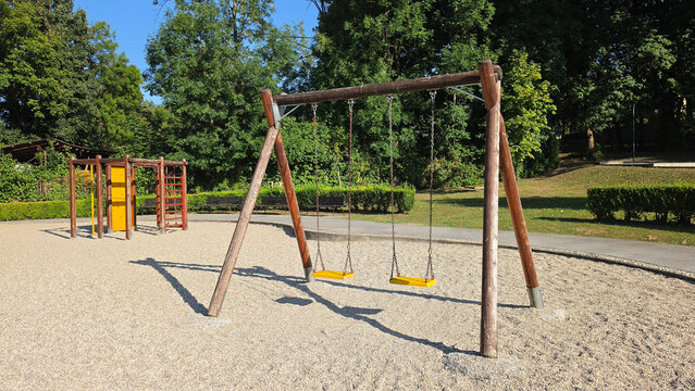 Wooden swing set with yellow seats standing on a gravel outdoor playground surrounded by green trees