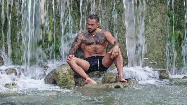 Man sitting on rocks beneath cascading water. Muscular man resting barefoot beside flowing water. Man with tattoo sleeves wearing black swimwear. Bearded man facing the camera among rocks.