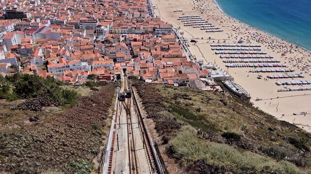 Two Nazar&eacute; funicular cars cross on the hillside, with one ascending while the other simultaneously travels in the opposite, descending direction toward the town and beach in Portugal.