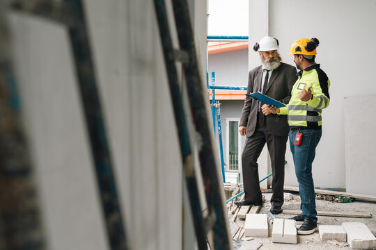 Construction worker and architect discuss building progress inside unfinished structure with safety equipment, blueprints, and construction materials, conveying focused professional collaboration
