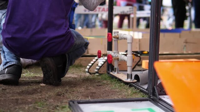 Students crouch on grassy ground while adjusting and testing compact tracked robot near plastic pipe obstacle setup in outdoor STEM robotics competition under soft daylight
