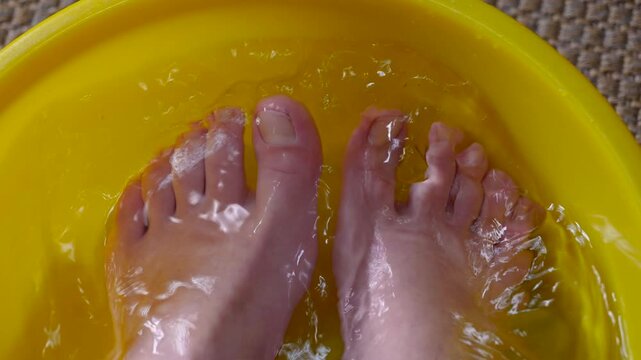 Person enjoying relaxing foot bath therapy. Close-up of bare feet soaking in a yellow basin of water, a relaxing therapeutic treatment to soothe tired limbs and promote hygiene after a day of work