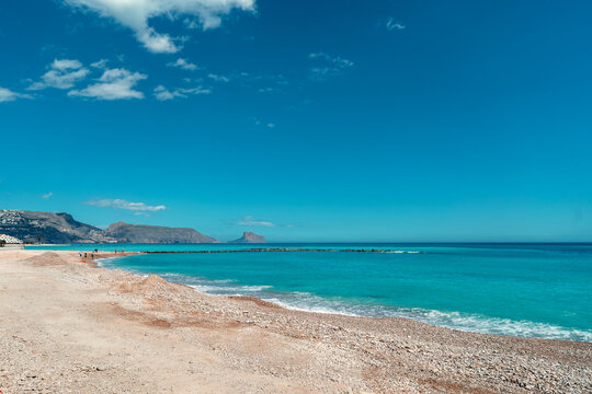 Pebble beach shoreline with clear turquoise Mediterranean sea water in Altea with Pe&ntilde;on de Ifach rock in distance, Alicante, Spain