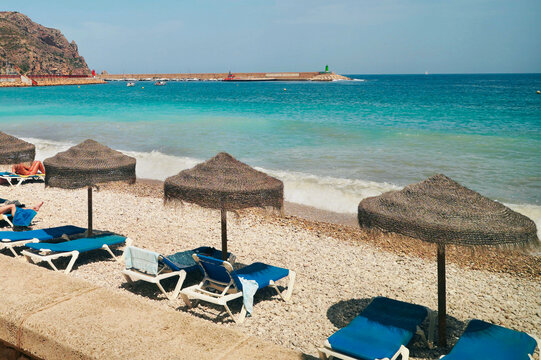 Straw beach umbrellas and blue sunbeds on pebble beach with sea view in Altea resort, Alicante, Spain