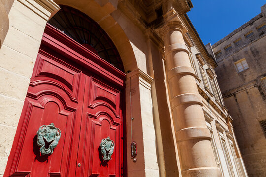 Grand arched red door with ornate bronze knockers set into a limestone facade with a decorative fluted column, in Rabat, Mdina, Malta