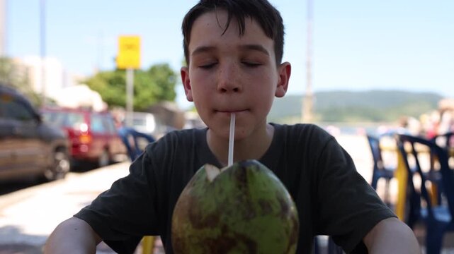 Boy drinking from fresh coconut beside beach in tropical city in summer