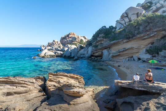 Two people relaxing beside a secluded rocky cove with clear turquoise water and sculpted granite formations at Capo Testa, northern Sardinia.