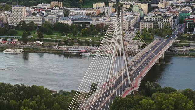 Smooth drone flight around Swietokrzyski Bridge over the Vistula River in Warsaw, aerial view of cable-stayed structure, moving traffic, waterfront, green parks and urban architecture