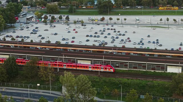 Passenger train departing from an urban railway station as drone smoothly flies alongside, aerial view of rail tracks, large parking area, buses, cars and surrounding transport infrastructure