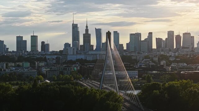 Drone flight from left to right across Warsaw at golden hour, revealing cable-stayed bridge, busy traffic, green parkland and panoramic downtown skyline with modern skyscrapers