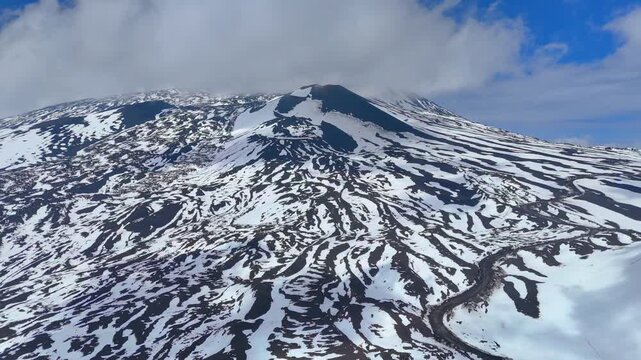 Drone aerial view of Mount Etna summit cone with snow patterns and winding mountain road in Sicily