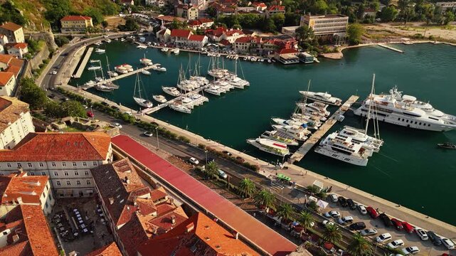 aerial tracking view of luxury motor yachts and sailing catamarans berthed at marina pontoons below the medieval town wall and promenade of kotor in low afternoon light, montenegro