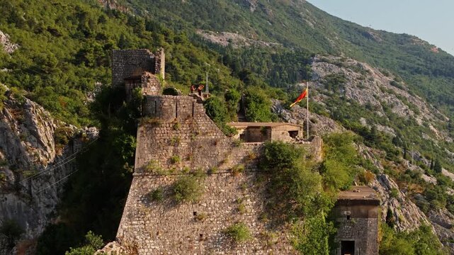 kotor, montenegro - aerial orbit around a ruined stone bastion of the kotor fortress with a montenegrin flag, green scrub and a forested ridge behind in soft afternoon light