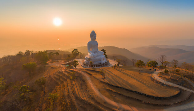 A large white Buddha statue sits atop a hill at sunrise, overlooking rolling mountains and casting long shadows across a serene landscape.