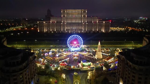 Aerial drone footage of the festive Bucharest Christmas Market illuminated at night in Constitution Square (Piata Constitutiei), featuring the glowing Ferris wheel, decorated Christmas tree.