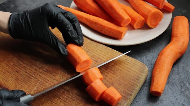 Gloved hands slice fresh peeled carrots on a wooden cutting board, preparing for cooking.