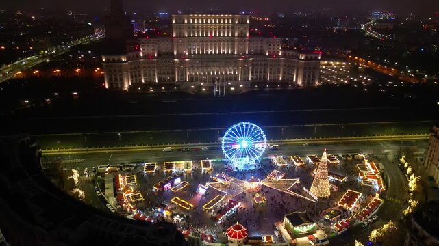 Aerial drone footage of the festive Bucharest Christmas Market illuminated at night in Constitution Square (Piata Constitutiei), featuring the glowing Ferris wheel, decorated Christmas tree.