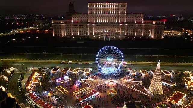 Aerial drone footage of the festive Bucharest Christmas Market illuminated at night in Constitution Square (Piata Constitutiei), featuring the glowing Ferris wheel, decorated Christmas tree.