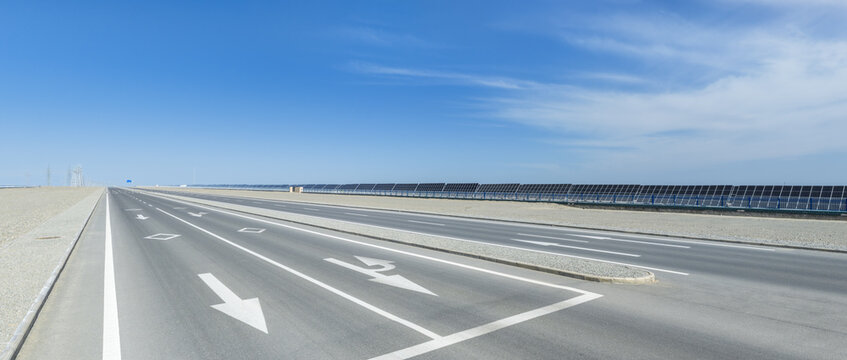 Empty Desert Highway with Lane Arrow Markings Running Beside Long Solar Photovoltaic Panel Arrays Under Blue Sky