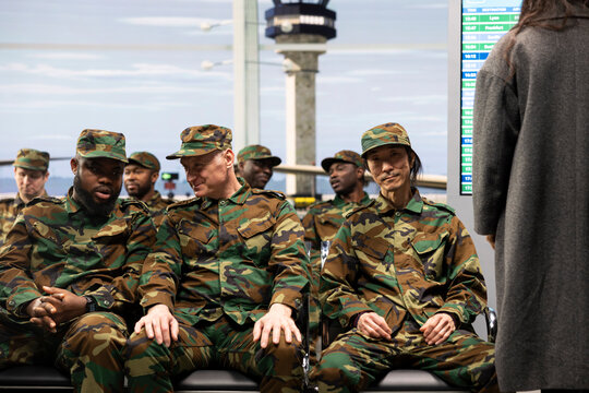 Patriotic army soldiers sit on an airport lounge bench and chat while waiting for their plane home, preparing for a homecoming reunion after a long deployment and spending time together.