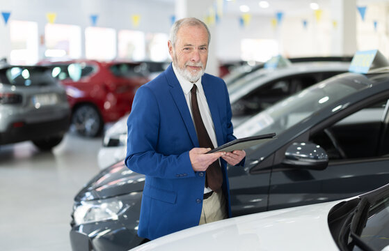 Elderly car salesman with tablet in car dealership offers various cars to potential buyers
