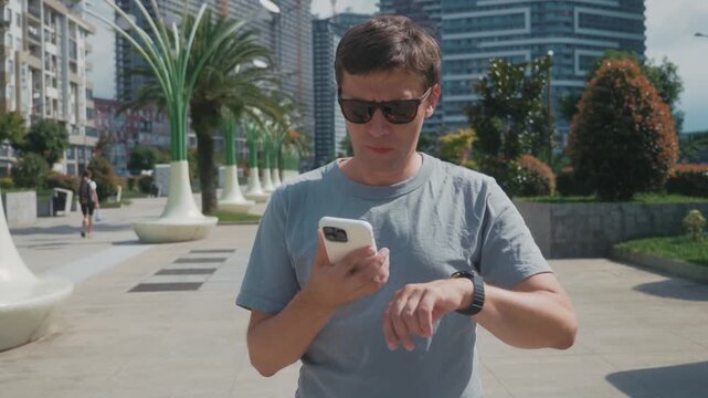 Man reviewing workout results in smartphone fitness app, tapping screen and checking training progress on smartwatch in urban park.