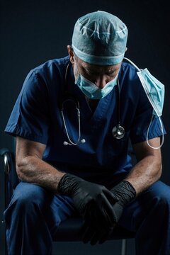 Doctor in blue scrubs sits with head down, exhausted after long night shift. Medical mask hangs off ear, stethoscope rests around neck. Black gloves on hands show fatigue of frontline worker.