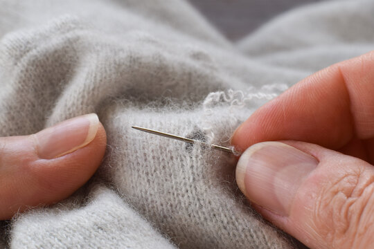 Close-up of hands darning and repairing hole in light-colored wool cashmere sweater using needle, showing sustainable clothing repair and textile maintenance