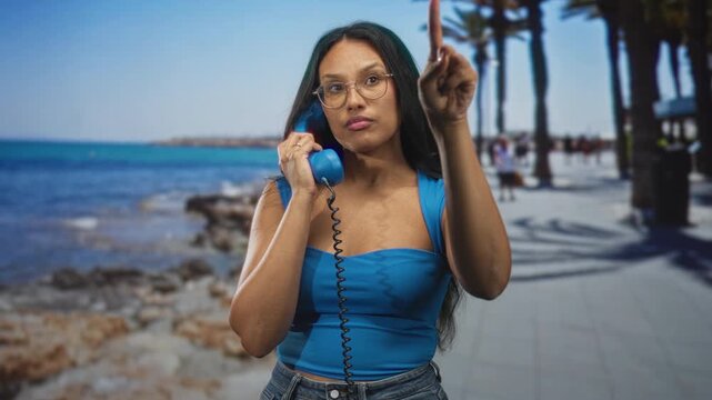 Woman wearing blue holding vintage telephone handset points finger on street promenade by ocean and palm trees; impatience.