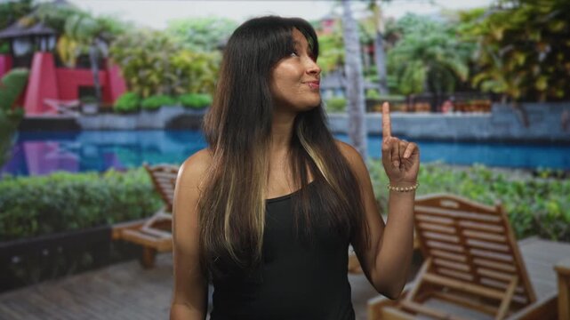 Woman points finger upward at hotel pool deck, smiling with eyes closed and long brunette hair in black tank top; serenity.