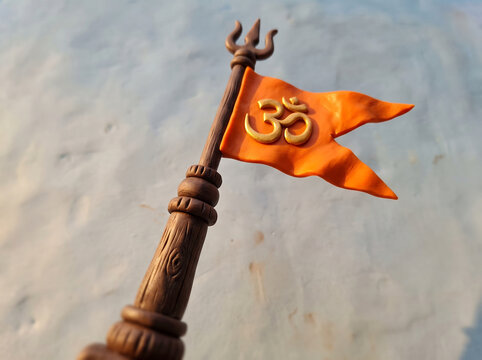 Miniature clay model of Hindu flag with Om symbol and Trishul trident on wooden staff against blurred background, Hindu festival or spirituality concept
