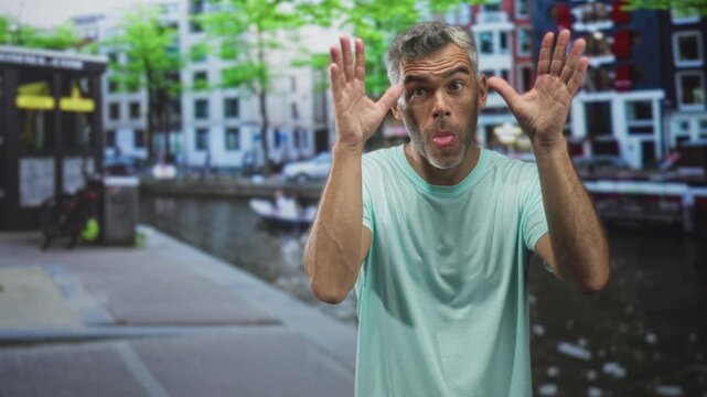 Man in green tshirt raises hands palms forward and sticks out tongue on a canal street; playful mischief.