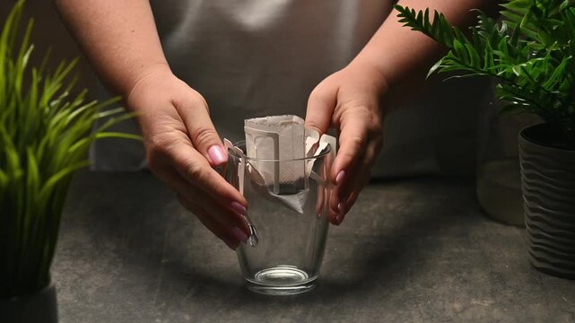 Hands fixing a single-serve drip coffee bag onto an empty glass cup on a stone counter. Static eye-level studio shot framed by potted plants with copy space