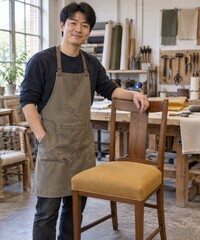 Fototapeta premium A young Chinese man apprentice stands beside his first completed ochre dining chair seat with a small satisfied smile and one hand resting on the wooden back in a bright upholstery training workshop