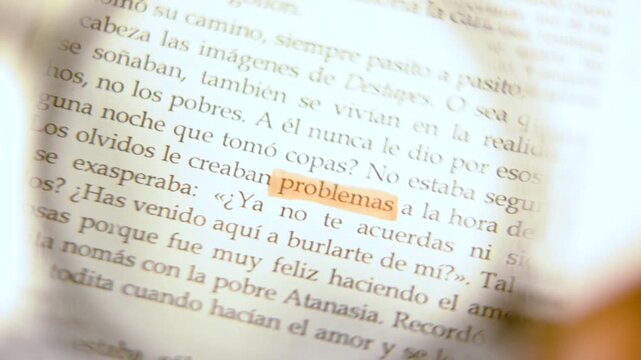4K slow-motion macro video of a magnifying glass zooming in on the orange highlighted word 'problemas' on a Spanish book page.