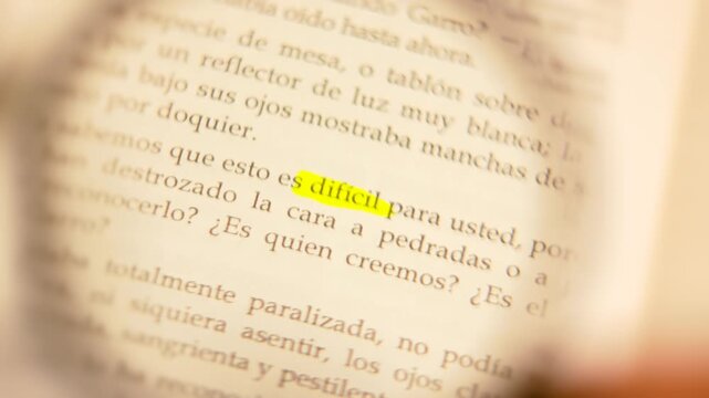 Macro close-up of a magnifying glass panning across a book page and bringing the highlighted word 'dificil' into sharp focus.