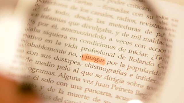 Cinematic 4K slow-motion macro shot of a magnifying glass enlarging the orange highlighted word 'juzgar' in a Spanish book.